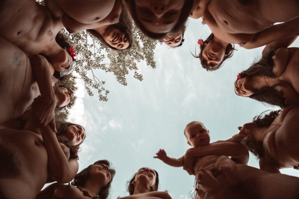 se ve un círculo de mujeres desnudas y abrazadas, alguna embarazada y otra sosteniendo a su bebé, sonriendo. La foto es tomada desde el suelo hacia el cielo.