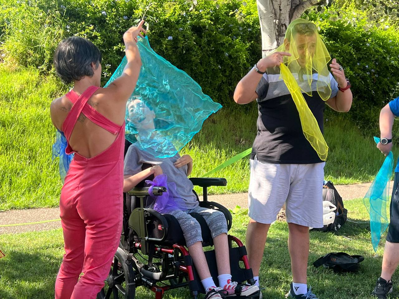 de espaldas muevo un pañuelo y se ve una mujer en silla de ruedas atras, en un circulo donde se ve un adulto con discapacidad haciendo malabares con los pañuelos y otro brazo de otra persona